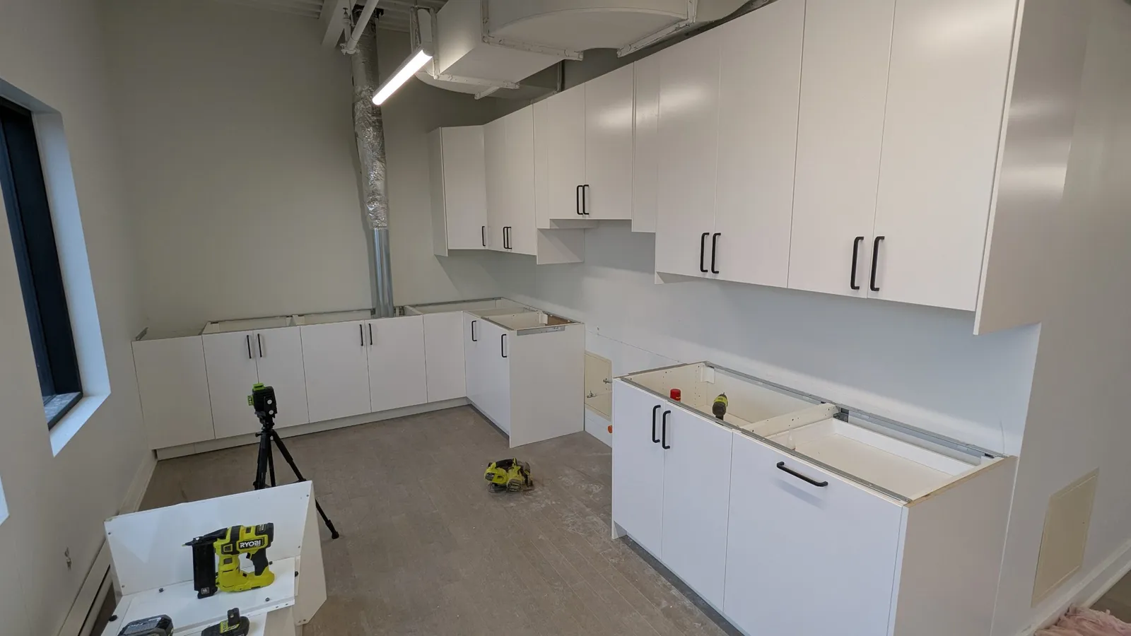 IKEA office kitchen in Montreal during installation, with white base and wall cabinets and black bar handles in an industrial space with exposed ductwork