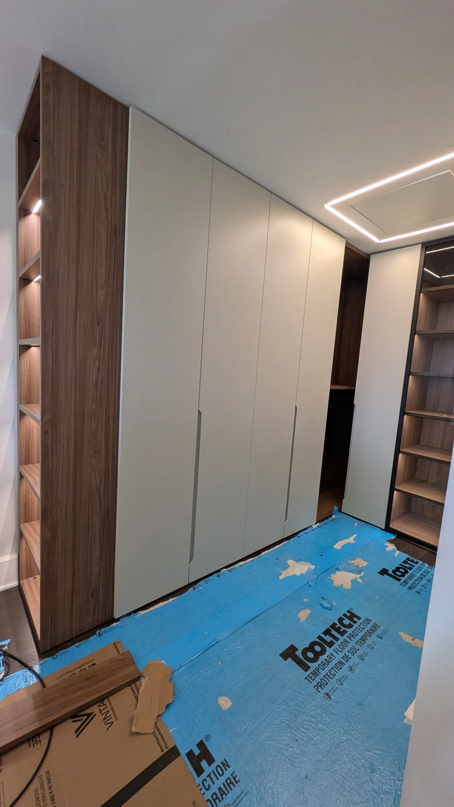 Custom floor-to-ceiling wardrobe with cream full-height doors and recessed handles beside a walnut open shelving unit with lit shelves