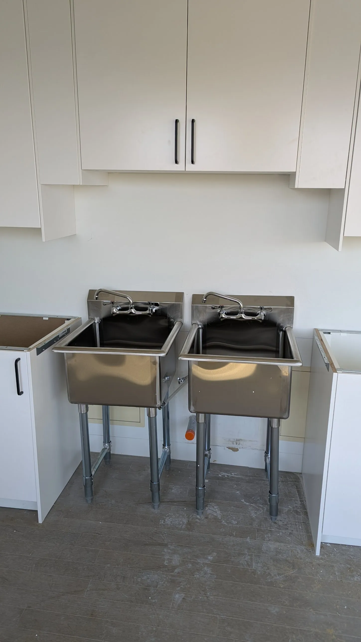 Two freestanding stainless-steel utility sinks between white IKEA base cabinets, with white wall cabinets above
