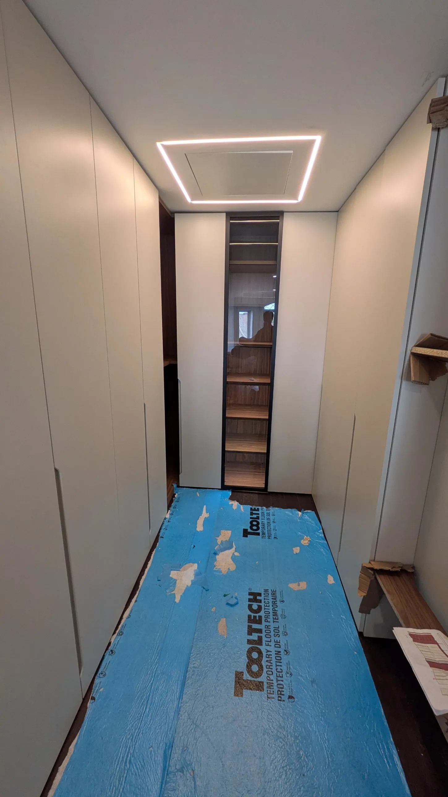 Walk-in closet corridor with full-height cream doors on both sides, a black-framed glass door with lit shelving and LED lighting around the ceiling panel